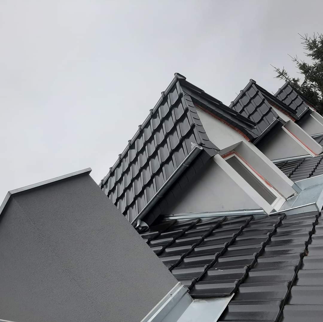 Fragment dachu z czarną dachówką karpiówką, lukarnami i orynnowaniem, sfotografowany w pochmurny dzień z dołu.