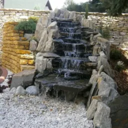 Kaskada wodna z kamieni łupanych na tle kamiennego muru, otoczona białym żwirem i roślinnością ozdobną.