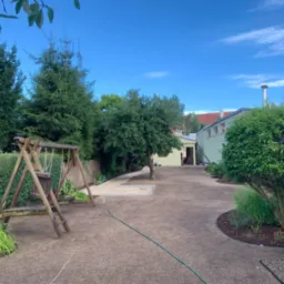 Przestronny ogród po wyrównaniu terenu, z przygotowanym podłożem pod trawnik, widoczne drzewa, krzewy, huśtawka i ścieżka z piasku.