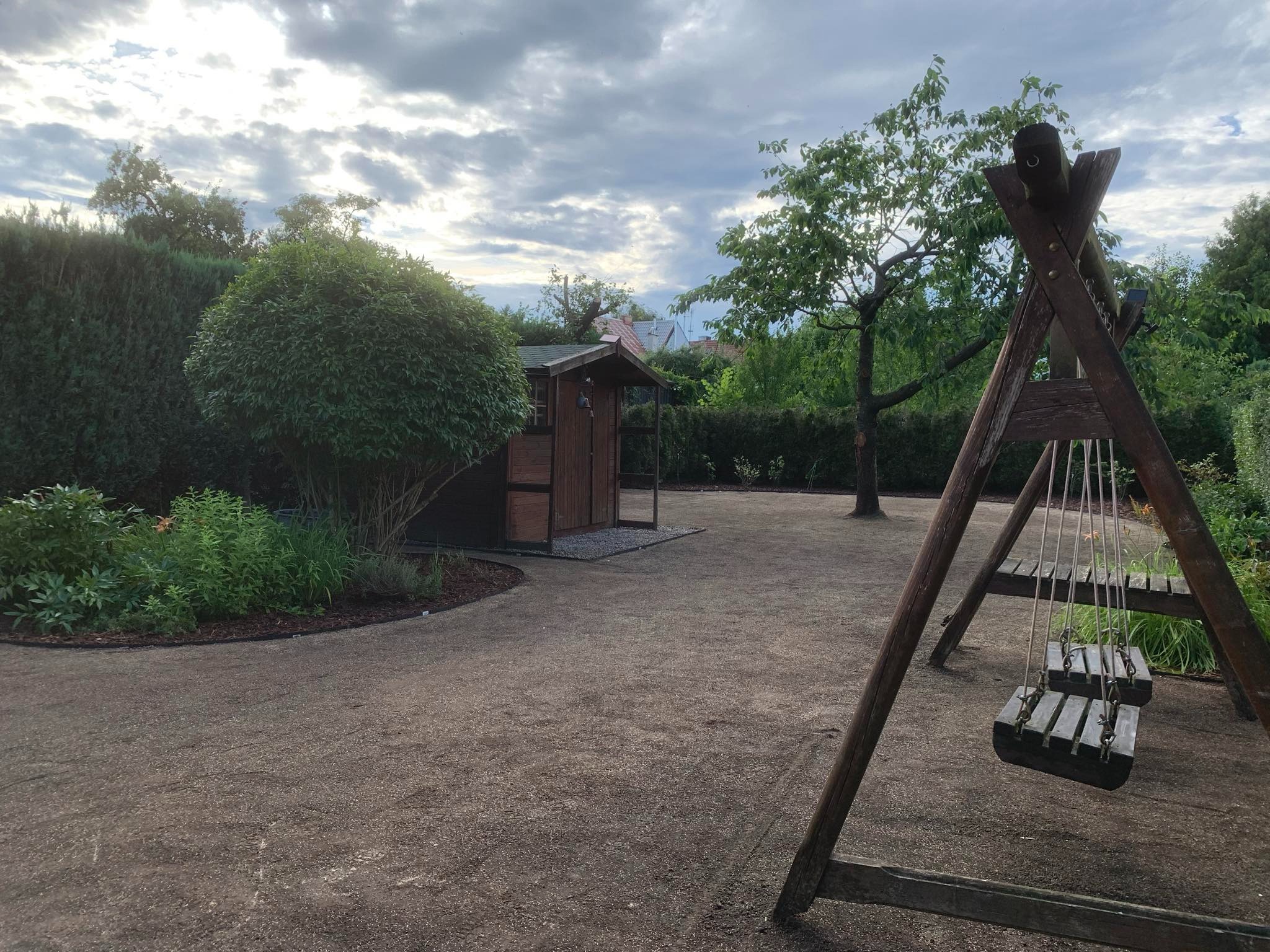 Uporządkowany ogród z drewnianym domkiem narzędziowym i huśtawką, świeżo wyrównany teren pokryty drobnym żwirem, otoczony zielenią i drzewami.