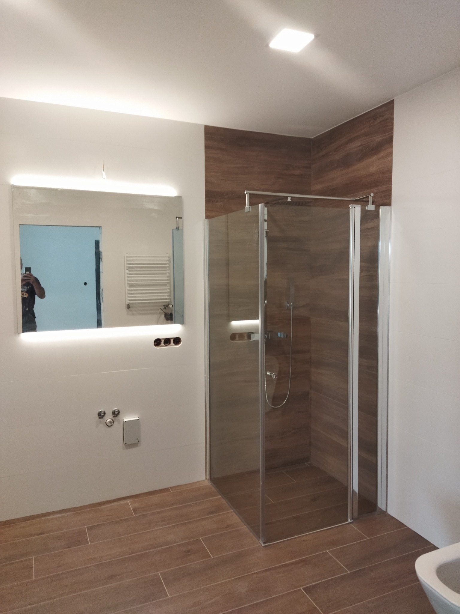 Nowoczesna łazienka po remoncie: kabina prysznicowa z brązowymi płytkami imitującymi drewno, podświetlane lustro z widocznym odbiciem grzejnika drabinkowego i osoby robiącej zdjęcie, oraz brązowa...