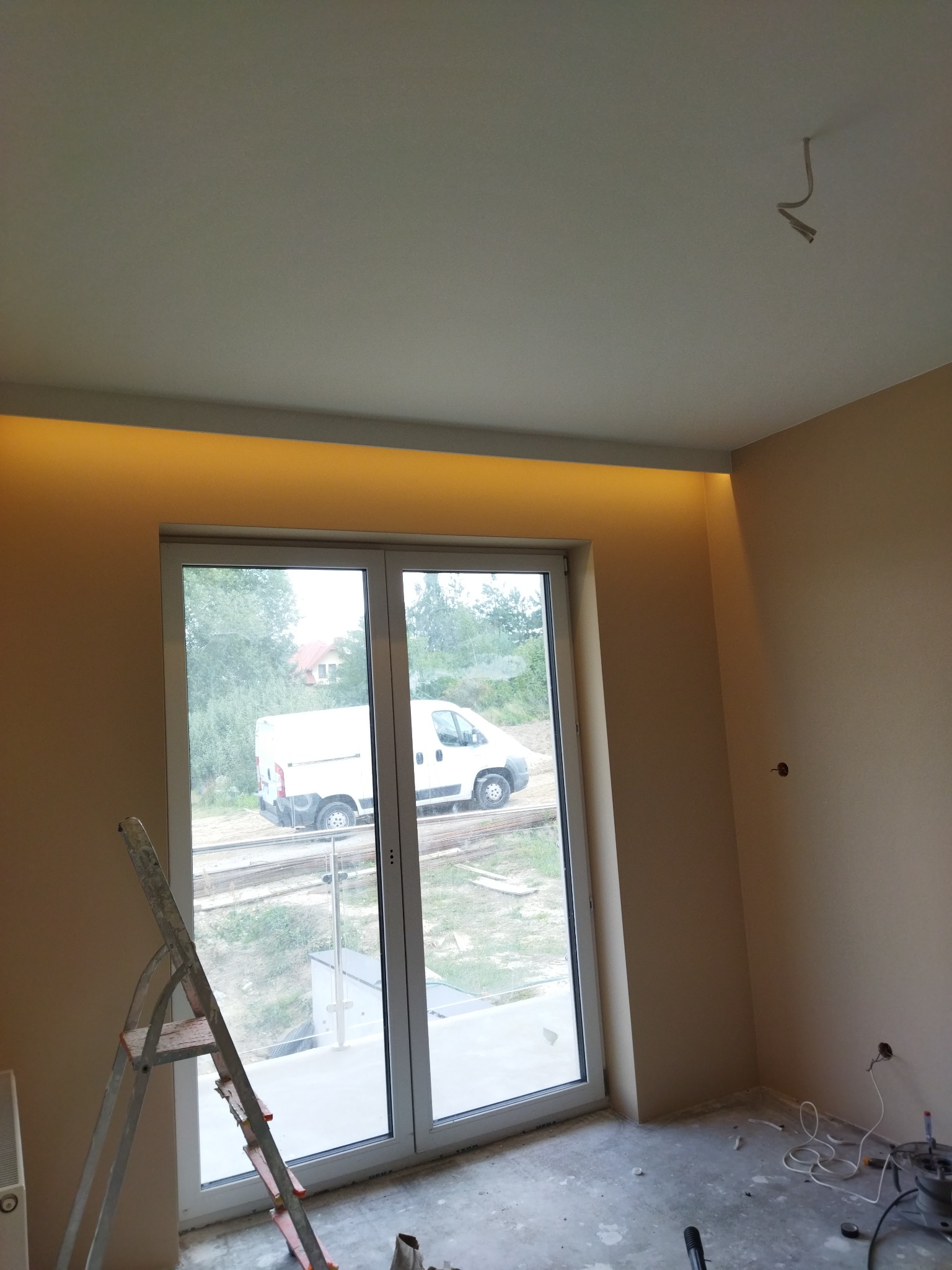 Pokój po szpachlowaniu i malowaniu ścian na beżowo, z oknem balkonowym i schodami w tle, widoczne przewody elektryczne wystające z sufitu i ściany oraz drabina oparta o okno.