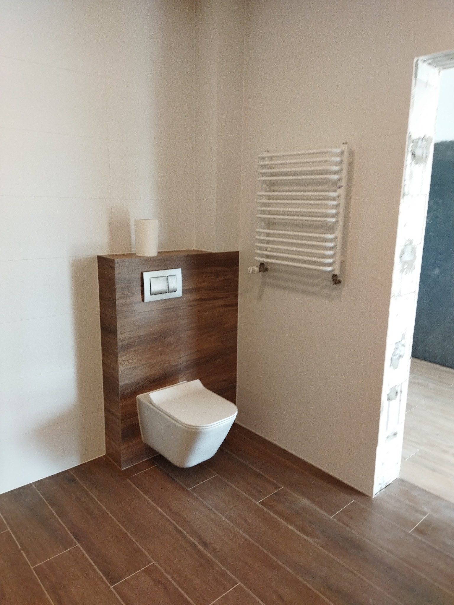 Nowoczesna łazienka z wiszącą toaletą, zabudową z drewnopodobnej płyty, grzejnikiem drabinkowym i brązowymi płytkami podłogowymi imitującymi drewno.