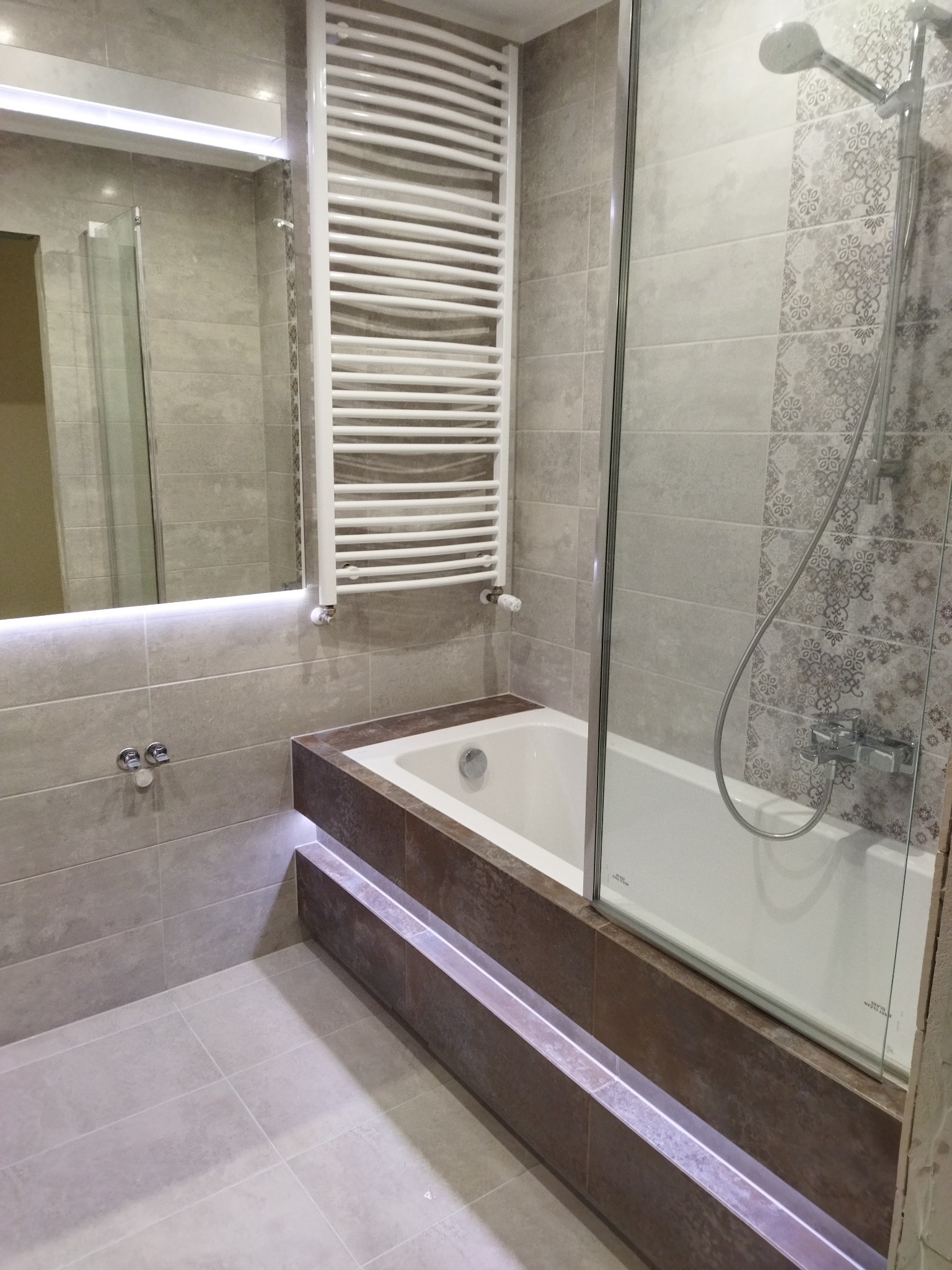 Wykończona łazienka w szarościach z podświetlaną wanną, grzejnikiem drabinkowym i kabiną prysznicową z dekoracyjnymi płytkami.