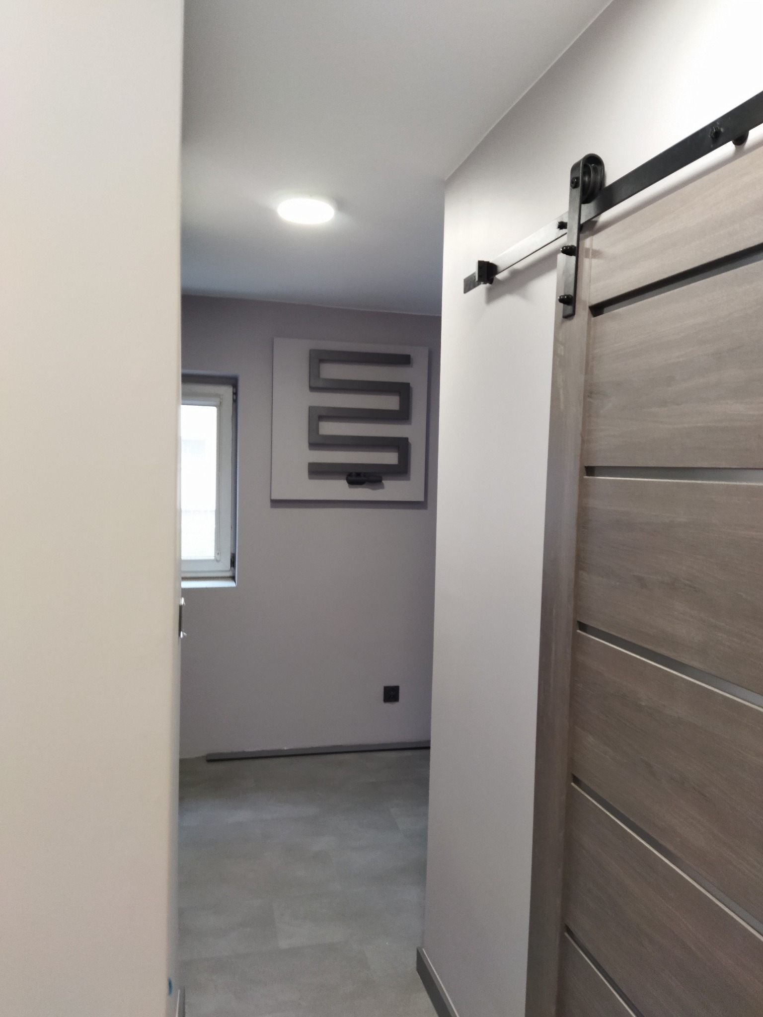 Szara, drewniana, przesuwna brama stodoły z czarnym osprzętem w nowocześnie odnowionym korytarzu z szarą podłogą i dekoracyjnym panelem ściennym.