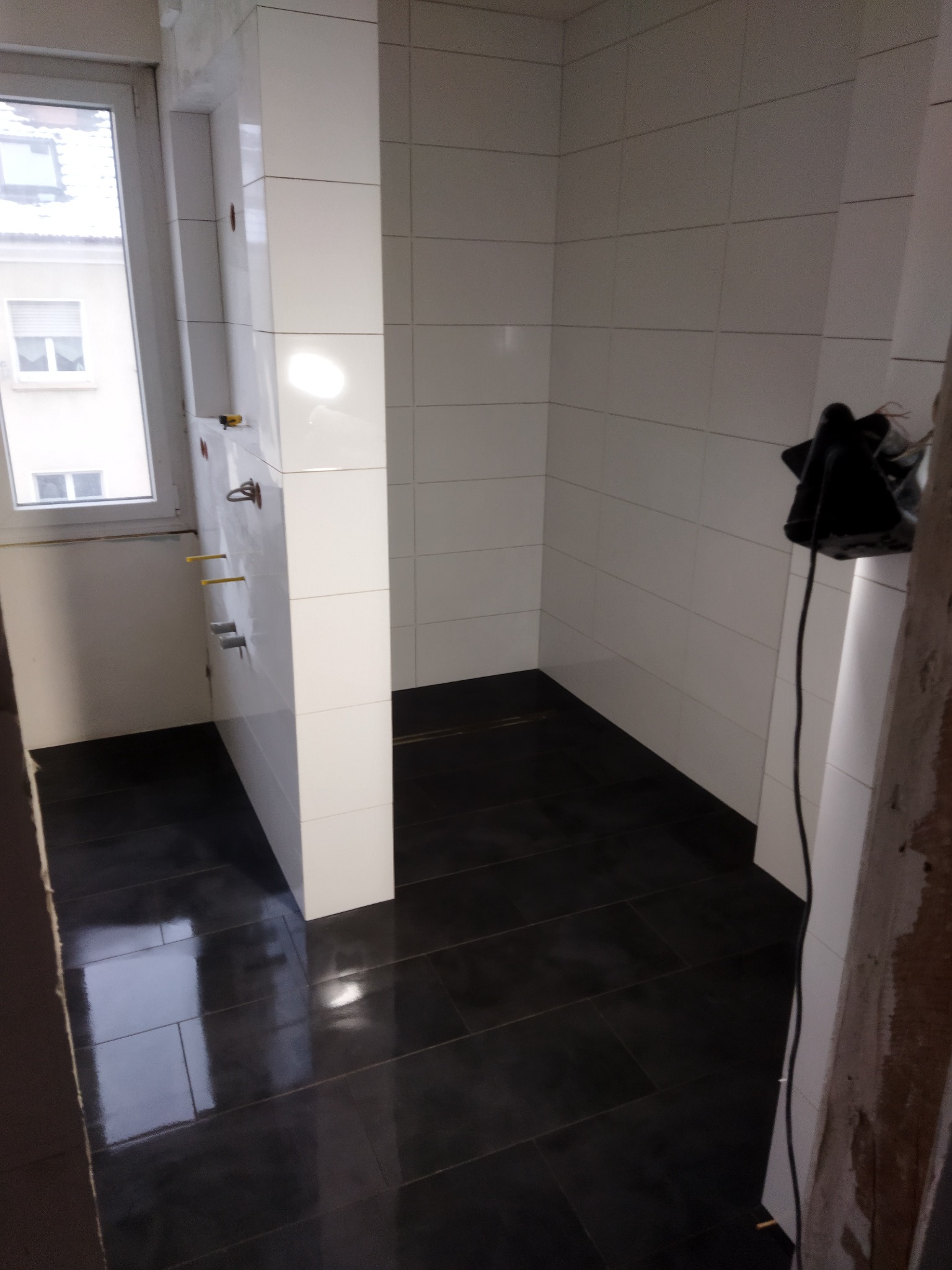 Wnętrze łazienki w trakcie remontu: białe płytki ścienne ułożone do połowy wysokości, ciemna, błyszcząca podłoga z płytek, wystające rury instalacji wodnej, widok na okno.