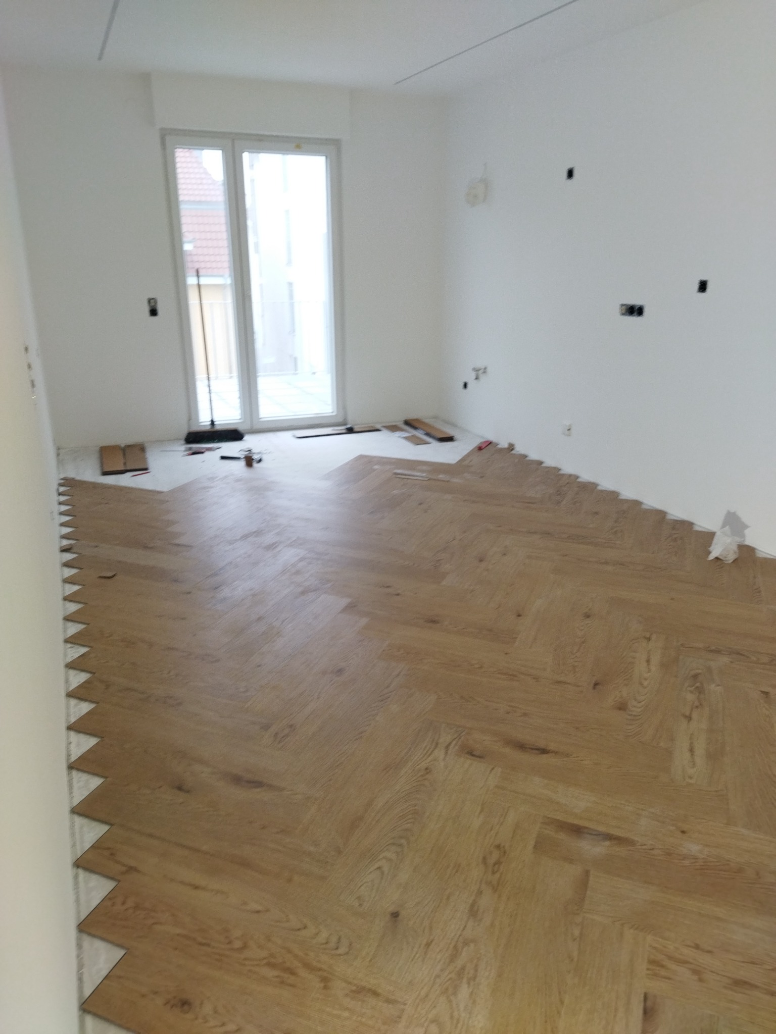 Układanie parkietu w jodełkę w jasnym, pustym pokoju z dużym oknem; widoczne narzędzia i niedokończone brzegi podłogi.