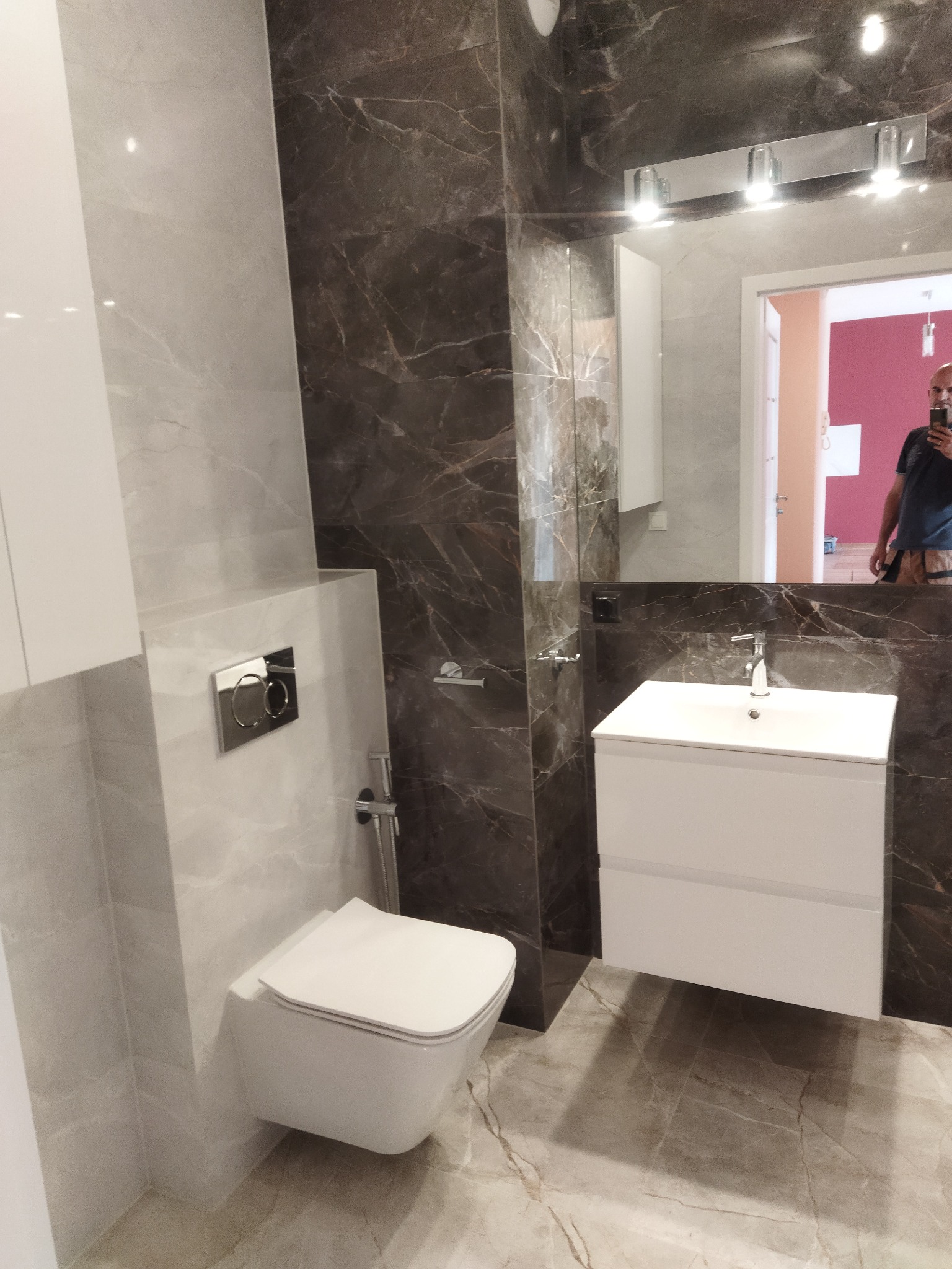 Nowoczesna łazienka z brązowo-szarymi płytkami imitującymi marmur, podwieszana toaleta z chromowanym przyciskiem spłukiwania, biała szafka z umywalką oraz lustro z oświetleniem. W odbiciu lustra...