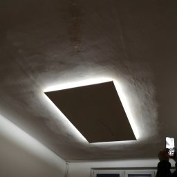 Łukasz Kostilek - Prostokątny panel sufitowy z podświetleniem LED, widoczny z dołu, z telefonem w dłoni osoby fotografującej w prawym dolnym rogu.