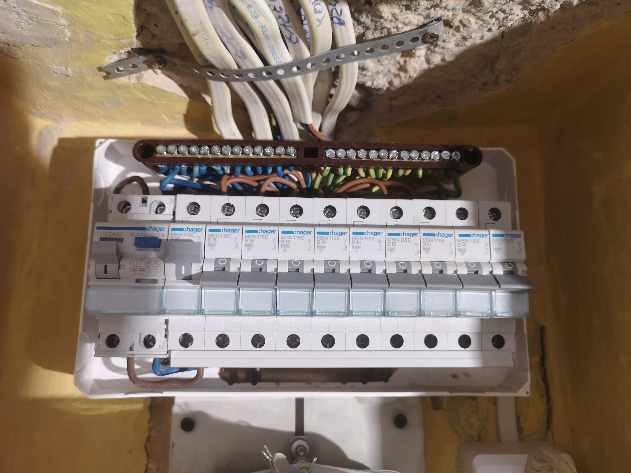 Rozdzielnica elektryczna Hager z podłączonymi przewodami w żółtej ścianie, widoczne zabezpieczenia i listwa zaciskowa.