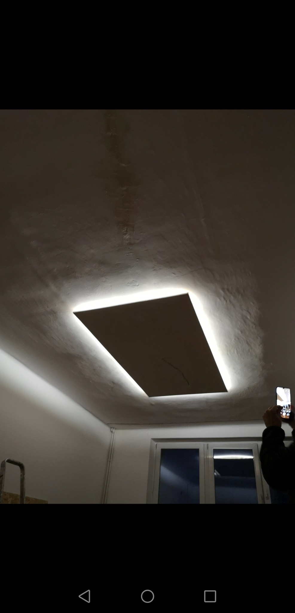 Prostokątny panel sufitowy z podświetleniem LED, widoczny z dołu, z telefonem w dłoni osoby fotografującej w prawym dolnym rogu.