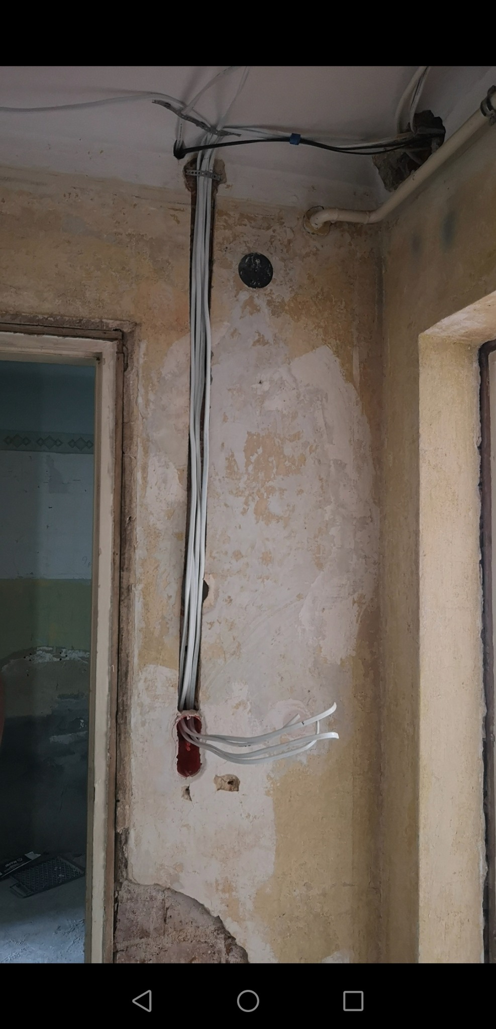 Pionowy układ kabli elektrycznych w ścianie z widoczną puszką instalacyjną i fragmentami starej farby, widok na sufit z przepustami kablowymi.