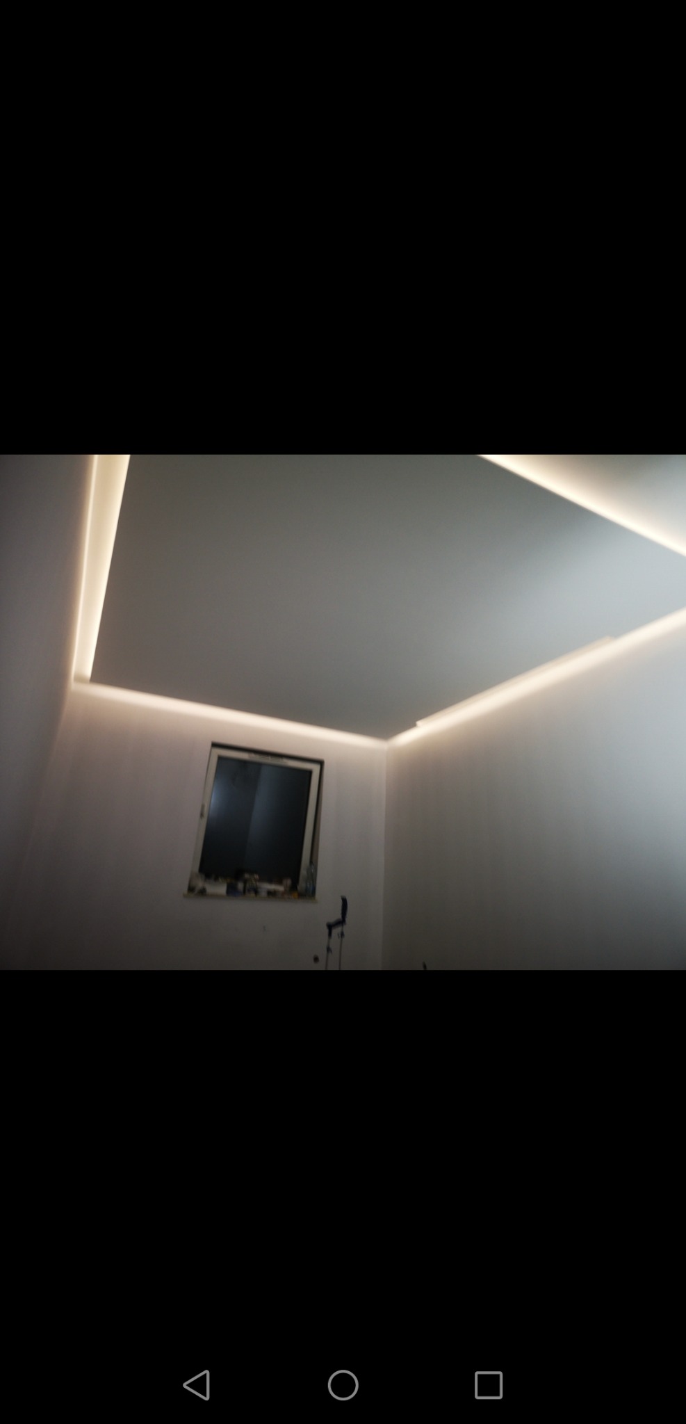 Pokój z białymi ścianami i sufitem, oświetlony listwami LED wzdłuż krawędzi sufitu, okno z widocznymi przedmiotami na parapecie. Widok z dołu.