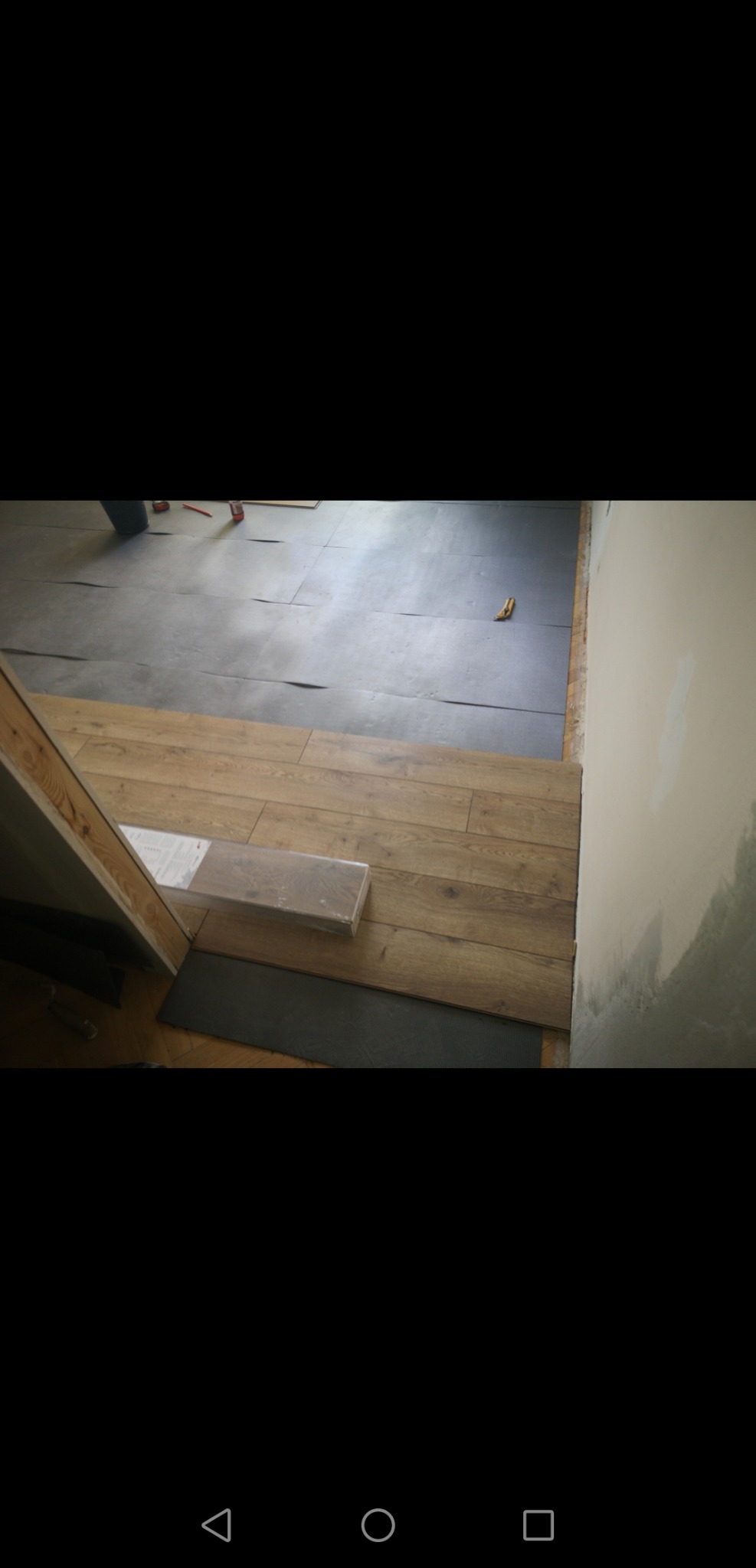 Widok na podłogę w trakcie remontu, gdzie część powierzchni pokryta jest szarą folią, a część panelami imitującymi drewno. Przy ścianie widoczne świeże ślady po malowaniu.