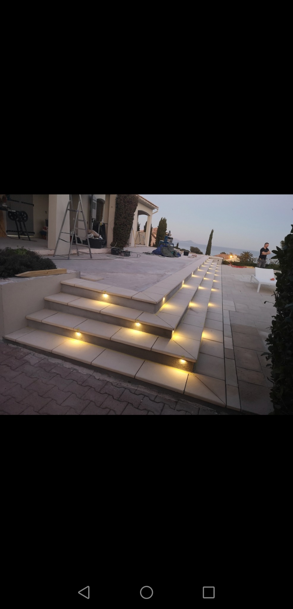 Schody zewnętrzne z jasnych płytek z oświetleniem punktowym w każdym stopniu, widok na dom w trakcie budowy i oddalony krajobraz morski o zmierzchu.