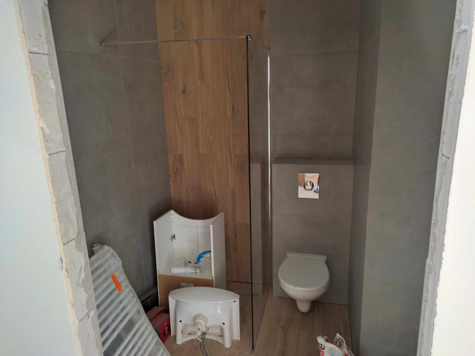 Niedokończona łazienka z drewnopodobnymi i szarymi płytkami, kabina prysznicowa ze szkła, wisząca toaleta i umywalka bez baterii, grzejnik drabinkowy oparty o ścianę.