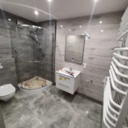 Nowoczesna łazienka w szarościach z kabiną prysznicową typu walk-in, umywalką na szafce i grzejnikiem drabinkowym, widoczne narzędzia i materiały wykończeniowe.