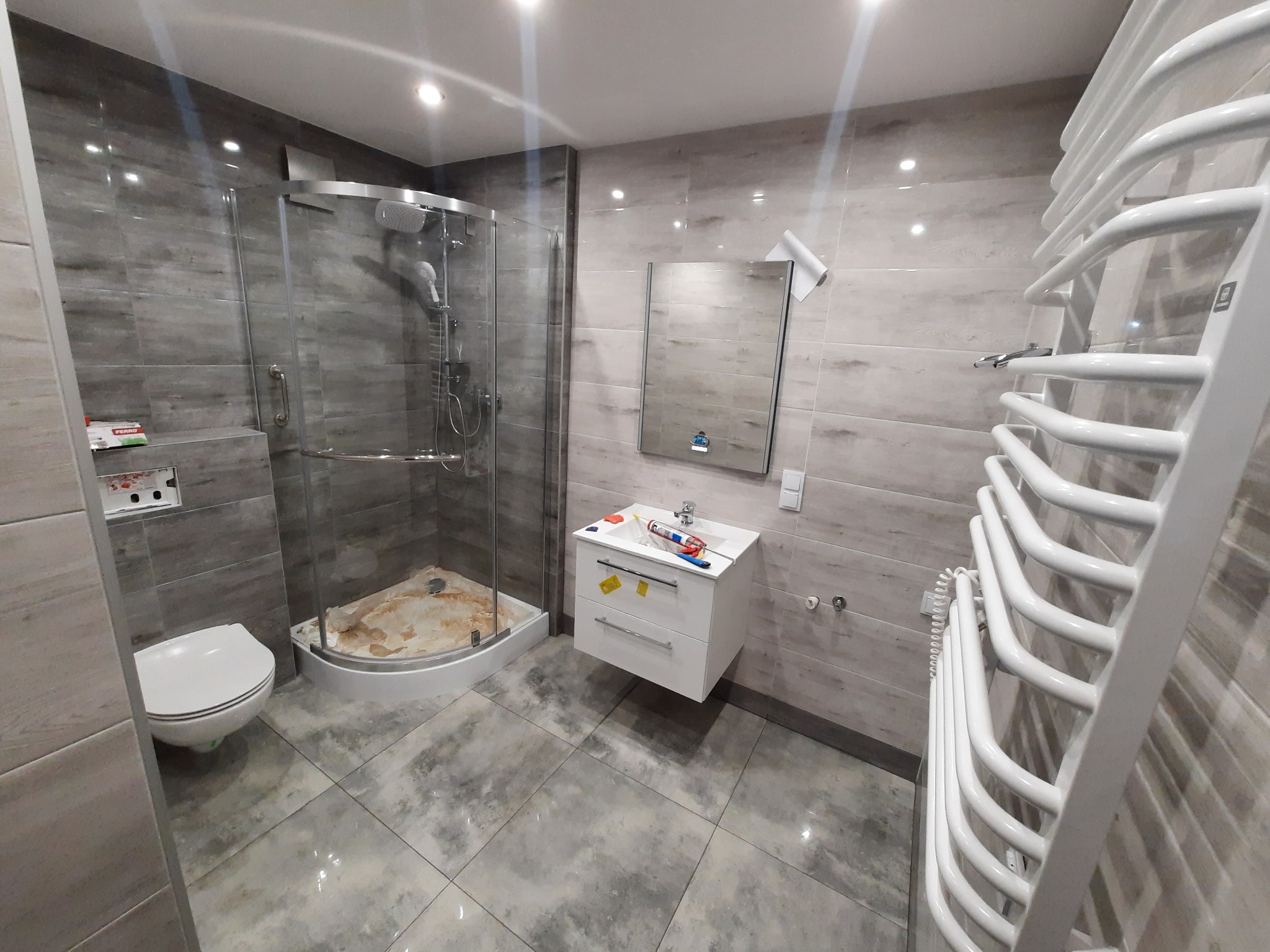 Nowoczesna łazienka w szarościach z kabiną prysznicową typu walk-in, umywalką na szafce i grzejnikiem drabinkowym, widoczne narzędzia i materiały wykończeniowe.