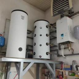 Dwa białe zbiorniki akumulacyjne na metalowej konstrukcji, z widocznymi przyłączami hydraulicznymi, obok zamontowany na ścianie biały moduł pompy ciepła Panasonic i grzejnik przemysłowy Euroheat...