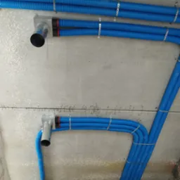 Niebieskie, karbowane rury wentylacyjne zamontowane pod szarym, betonowym sufitem za pomocą metalowych uchwytów, widoczne fragmenty instalacji z czerwonymi elementami.