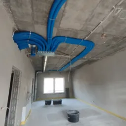 Niebieskie, karbowane rury wentylacyjne zamontowane pod surowym, betonowym sufitem w surowym wnętrzu. Widoczne elementy montażowe, metalowe skrzynki rozdzielcze i fragmenty instalacji elektrycznej.