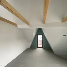 Poddasze po malowaniu ścian na biało, z drewnianymi belkami stropowymi i szarą wylewką podłogową, widok na drzwi balkonowe w szczycie ściany pomalowanej na zielono.