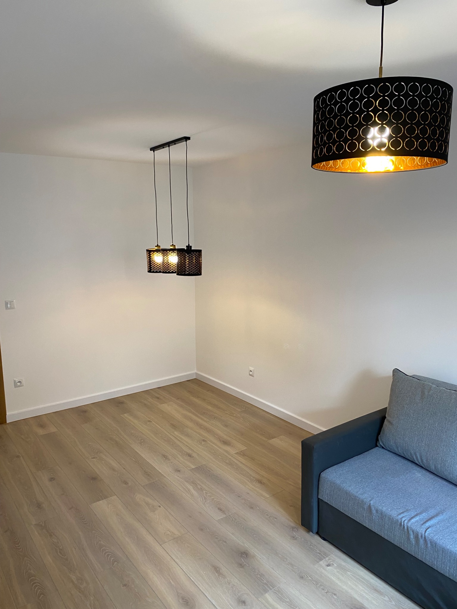 Jasne panele podłogowe w nowocześnie urządzonym pokoju z białymi ścianami, oświetlonym przez dwie czarne, ażurowe lampy wiszące, z szarą sofą w rogu.