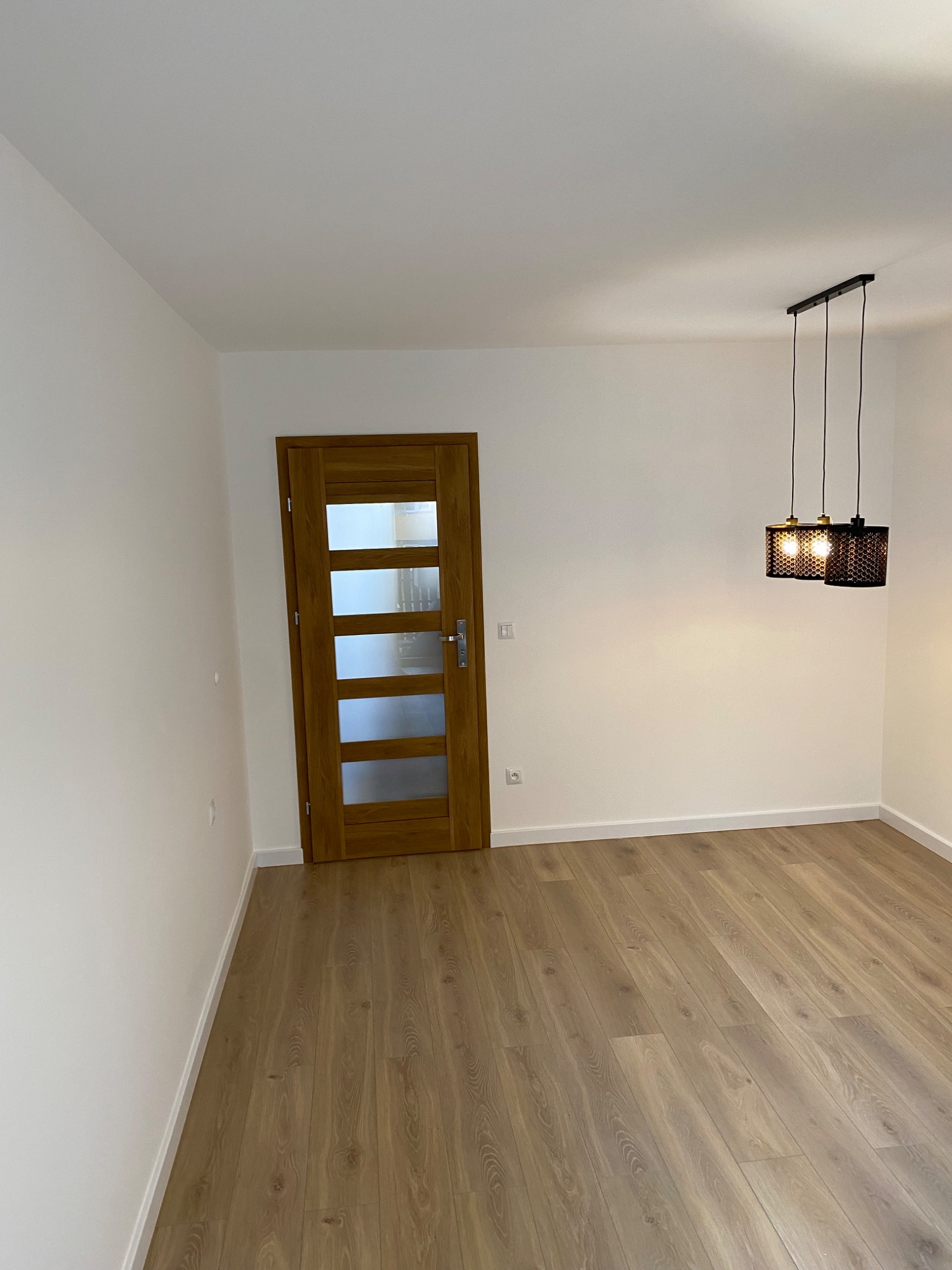Pusty pokój po remoncie z drewnianą podłogą, drzwiami z przeszkleniami i nowoczesną, metalową lampą wiszącą.