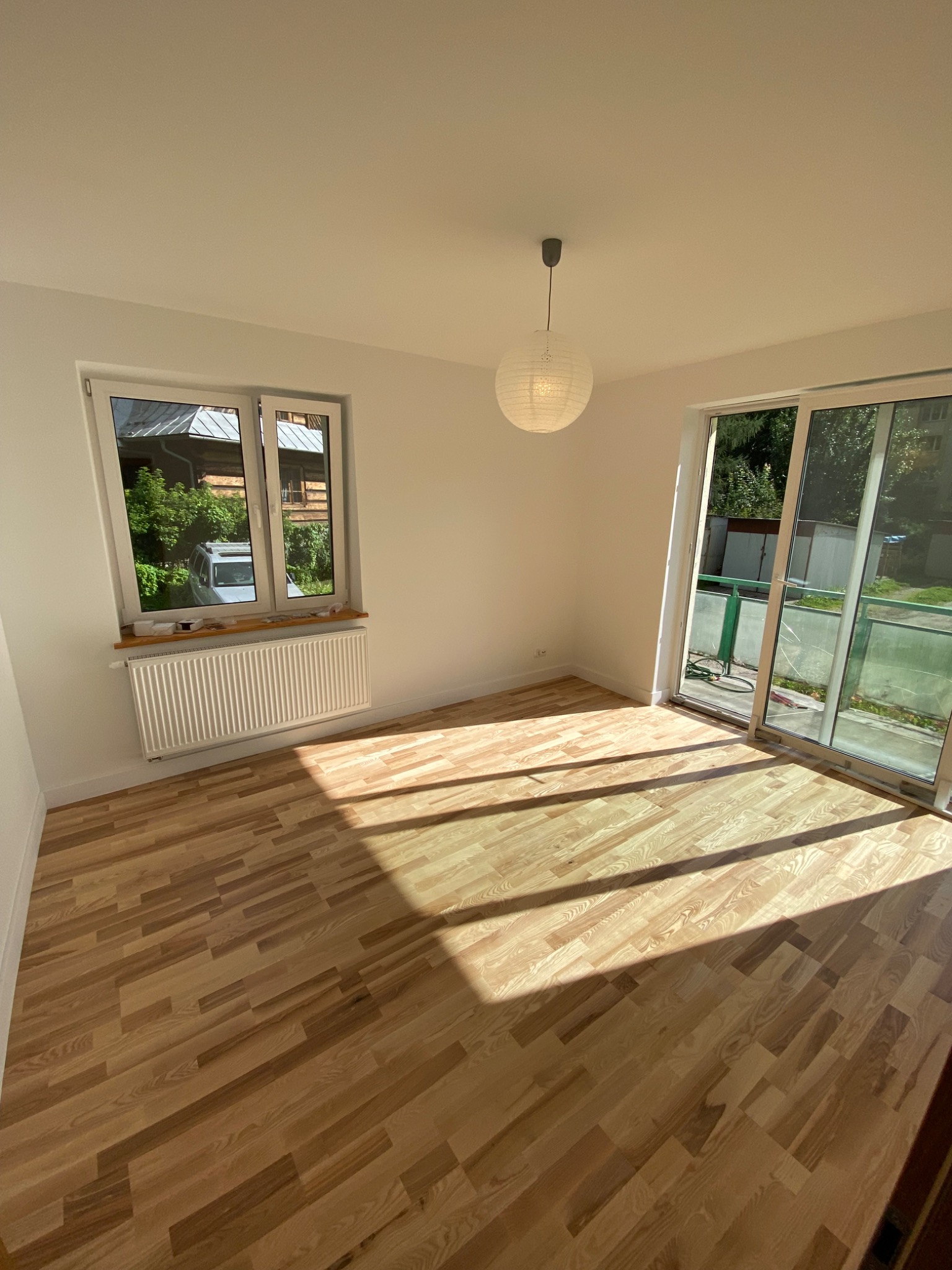 Jasne, puste pomieszczenie z nową drewnianą podłogą ułożoną we wzór parkietu, białymi ścianami, grzejnikiem pod oknem i lampionem wiszącym z sufitu, z balkonem widocznym przez przesuwne drzwi.