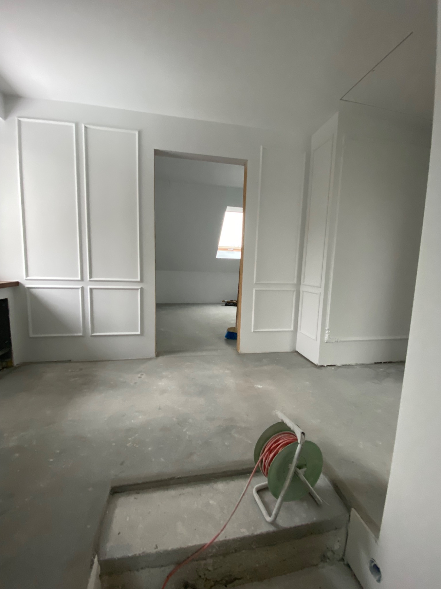 Wnętrze w trakcie prac wykończeniowych: surowa podłoga, białe ściany z dekoracyjnymi panelami, widoczny kabel na szpuli i przejście do kolejnego pomieszczenia z oknem dachowym.
