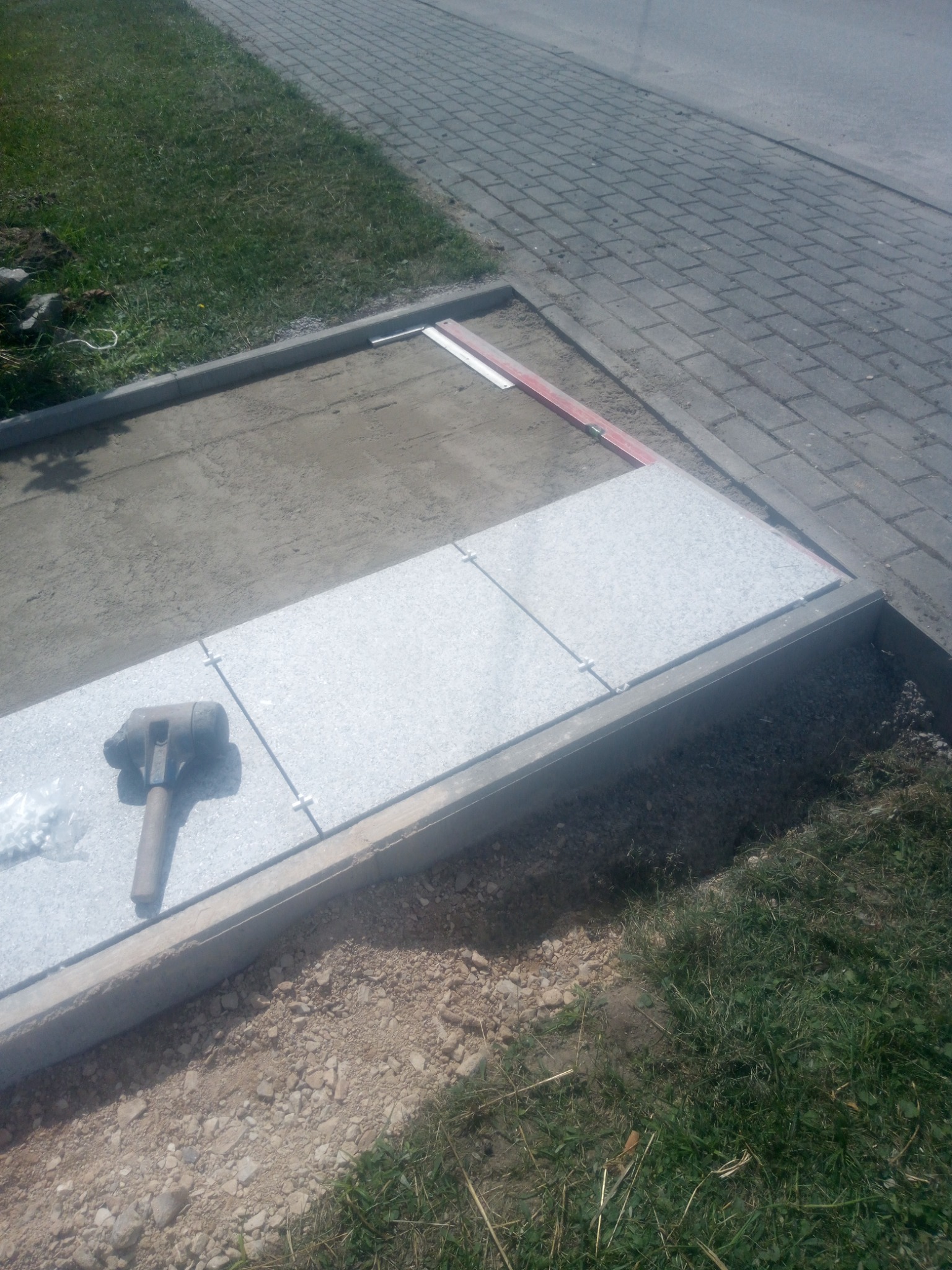 Montaż płyt granitowych w obramowaniu betonowym; widoczne narzędzia, poziomica i krzyżyki dystansowe.