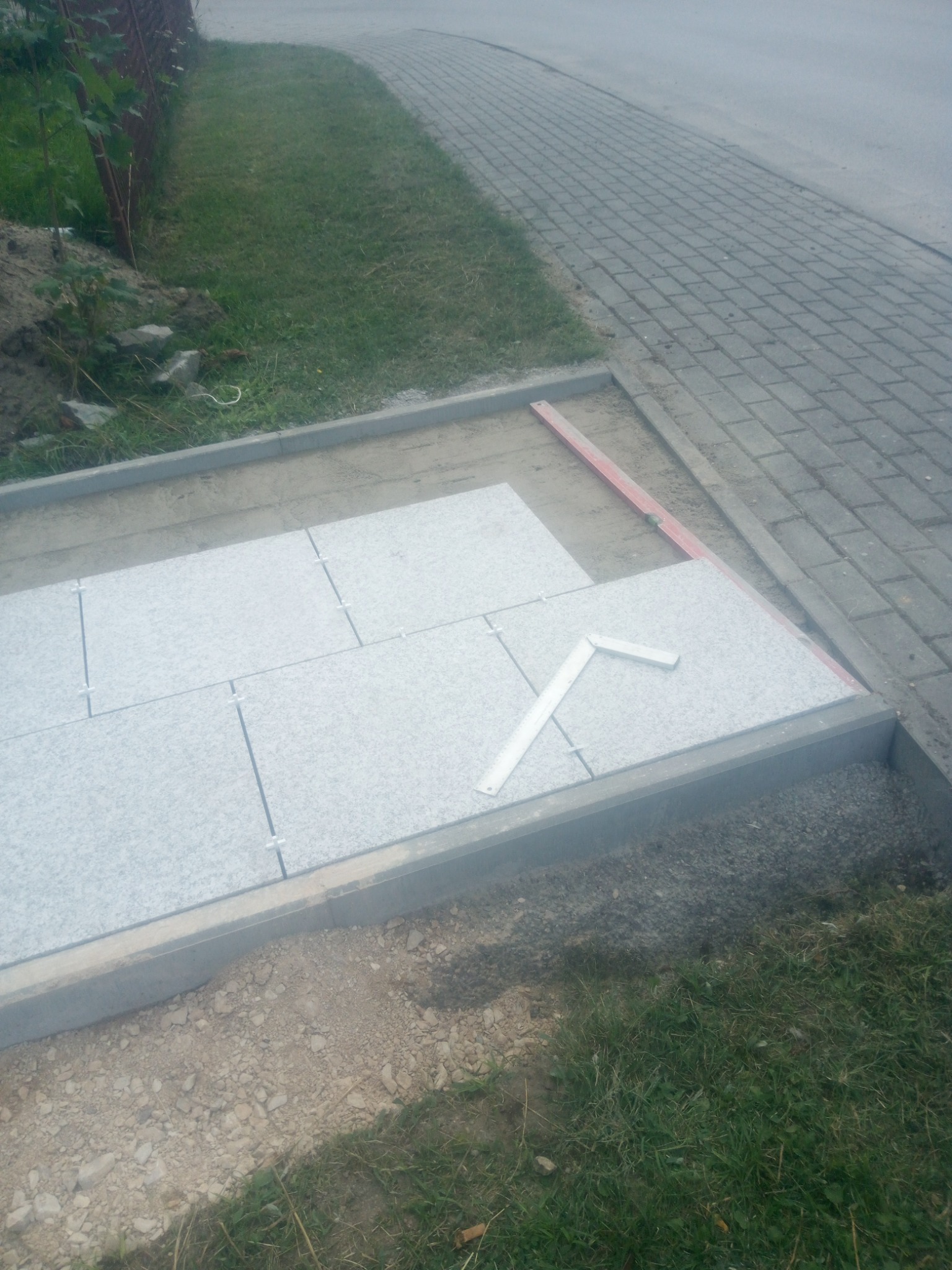 Fragment chodnika z szarych płyt granitowych w trakcie układania, widoczne krzyżyki dystansowe, poziomica i kątownik, obrzeże z kostki betonowej, trawa i kostka brukowa w tle.
