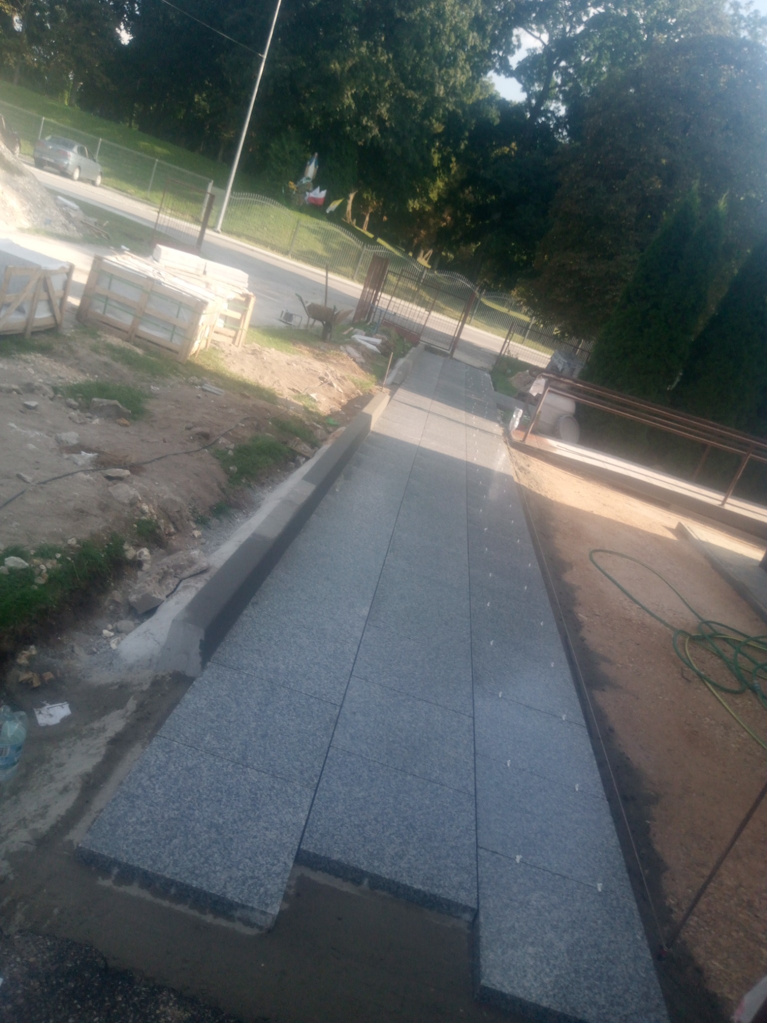 Fragment ścieżki z szarej kostki granitowej w trakcie budowy, widoczne palety z kostką w tle, ziemia i trawa po bokach, w oddali ulica i drzewa.