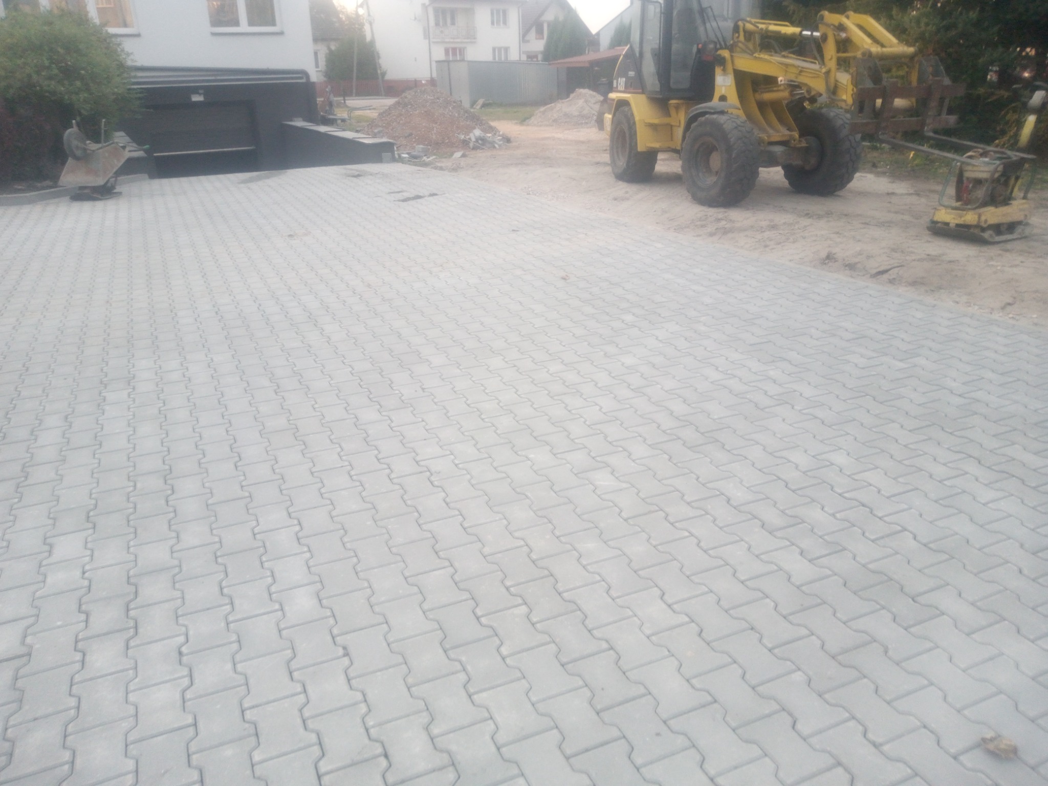 Ułożona szara kostka brukowa typu 'I' na podjeździe, w tle żółta ładowarka i ubijarka wibracyjna, widoczny budynek z garażem.