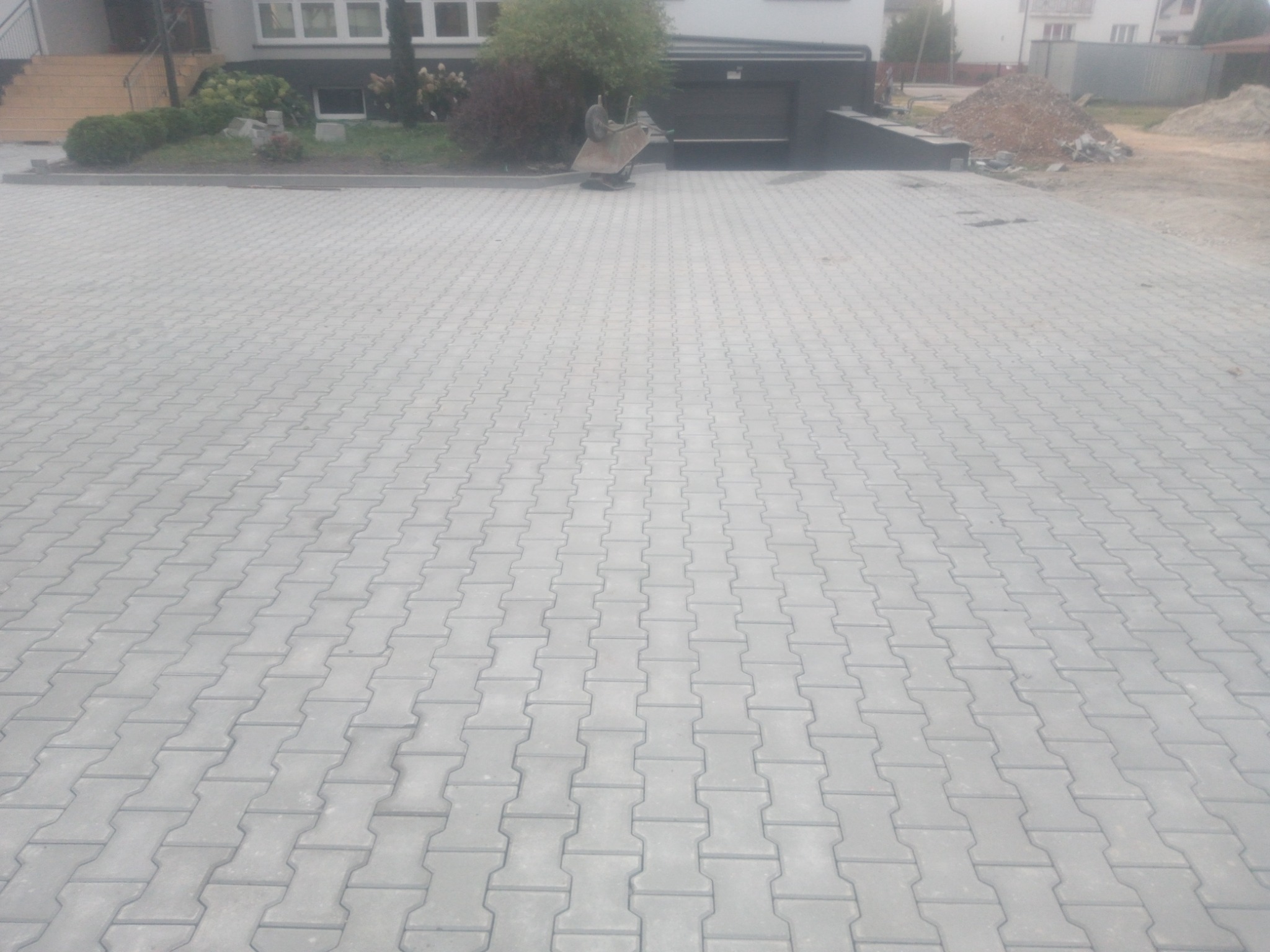 Ułożona, szara kostka brukowa w kształcie litery H na podjeździe przed domem z wózkiem budowlanym w tle.