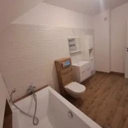 Nowoczesna łazienka na poddaszu z białymi płytkami imitującymi cegłę, drewnianą obudową toalety z przyciskami, białą wanną, umywalką z szafką i drewnopodobną podłogą.