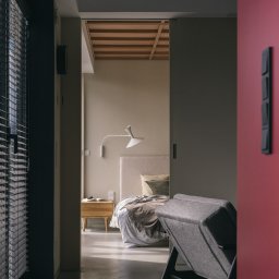 Marta Fibich - Nowoczesna sypialnia z widokiem na łóżko z drewnianym stolikiem nocnym i lampą, widziana zza szarego fotela i czerwonej ściany. Minimalistyczny design, stonowana kolorystyka.