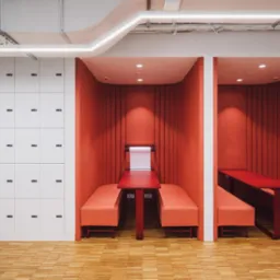 Nowoczesne biuro z dwoma pomarańczowymi kabinami do pracy, wyposażonymi w stoły i siedzenia, oraz białą ścianą szafek z numerowanymi zamkami, oświetlone ledowymi lampami sufitowymi.