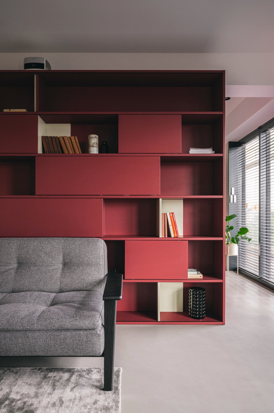 Nowoczesny salon z burgundową, modułową biblioteczką, szarą sofą i dywanem. Minimalistyczne wnętrze z rośliną w tle i żaluzjami.