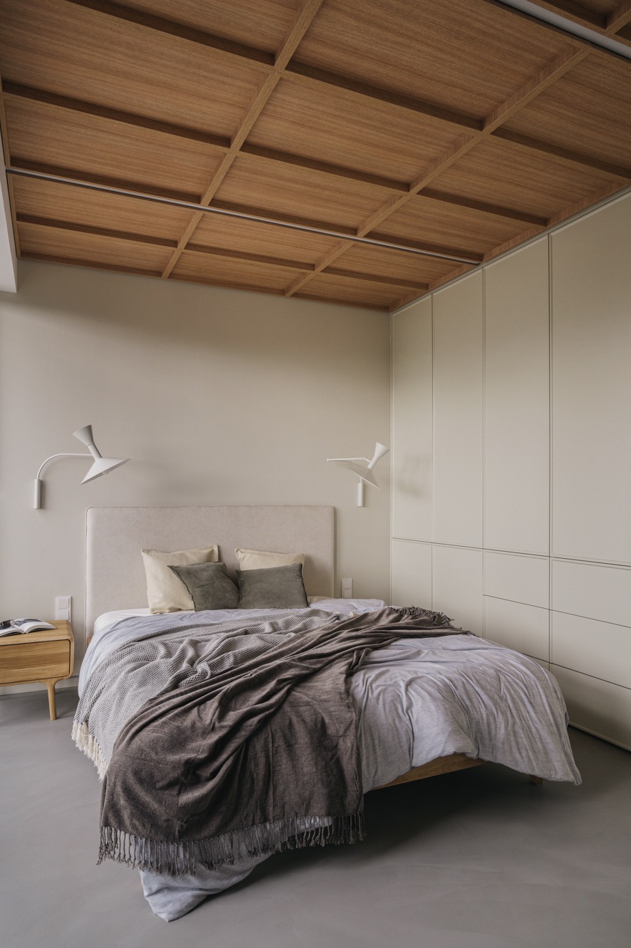 Minimalistyczna sypialnia z drewnianym sufitem kasetonowym, beżowymi ścianami, wbudowaną szafą i łóżkiem z narzutą w odcieniach szarości. Lampki nocne w stylu retro.