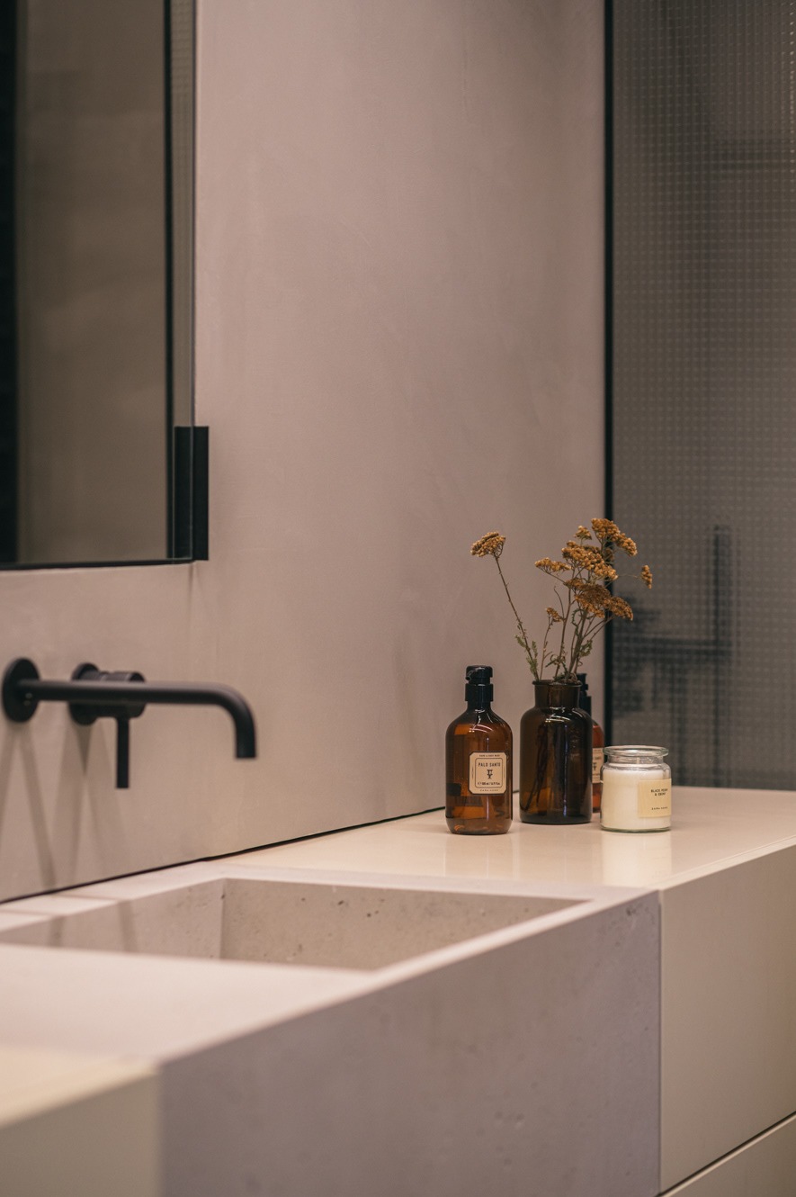 Minimalistyczna łazienka: betonowa umywalka z czarną armaturą, flakoniki i suszone kwiaty na blacie. Ściana w odcieniach szarości, lustro z czarną ramą.