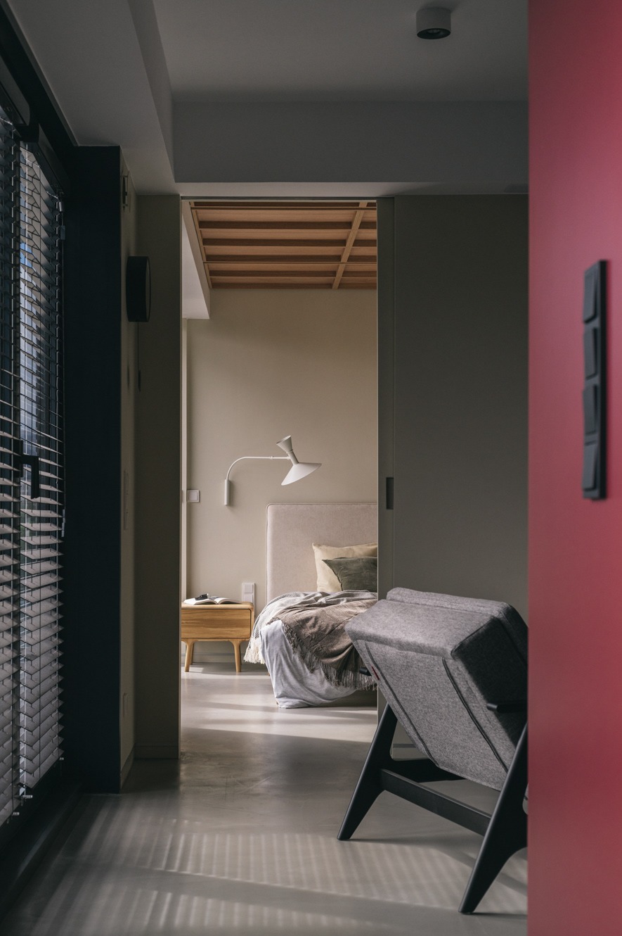 Nowoczesna sypialnia z widokiem na łóżko z drewnianym stolikiem nocnym i lampą, widziana zza szarego fotela i czerwonej ściany. Minimalistyczny design, stonowana kolorystyka.