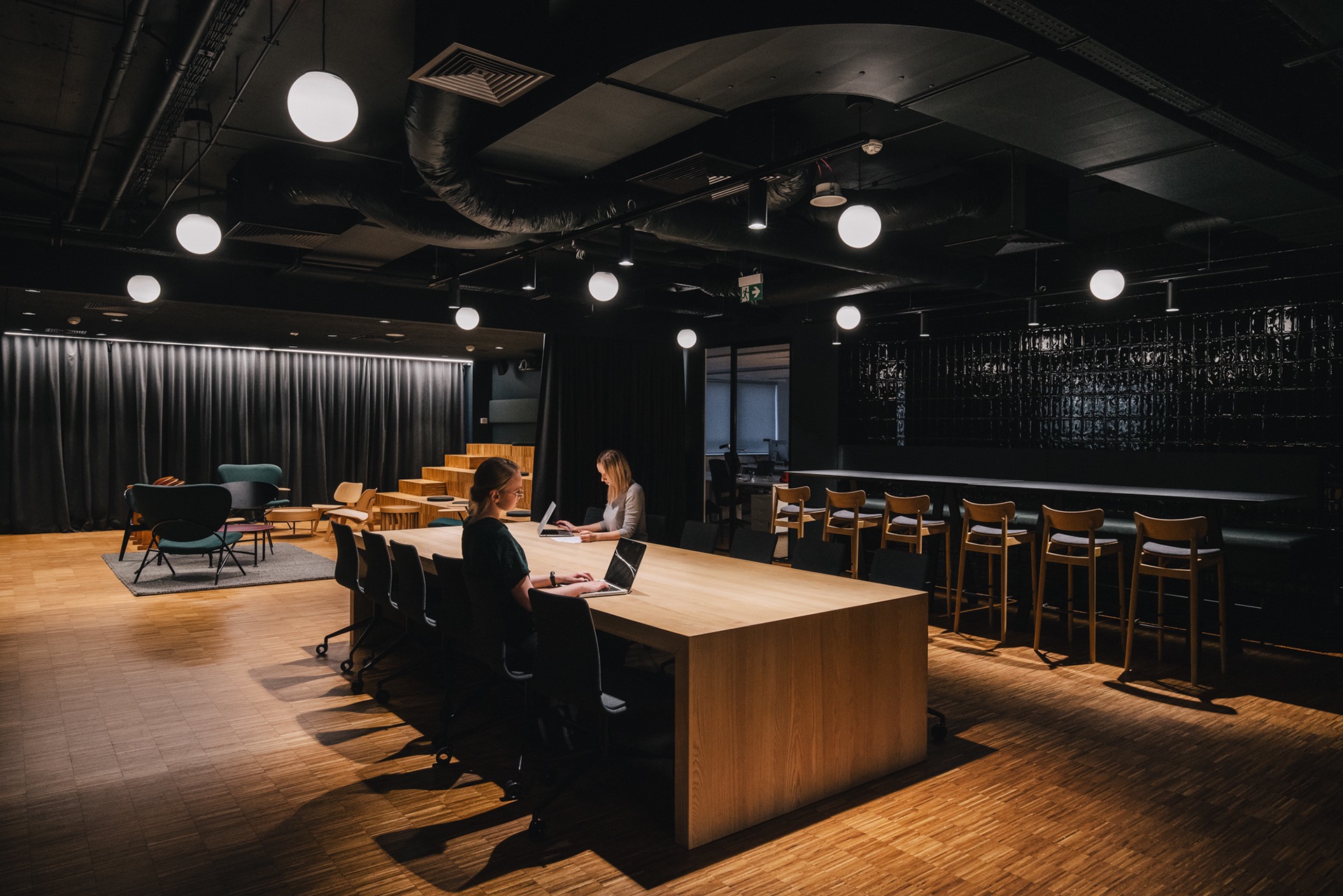 Przestronne biuro z drewnianą podłogą i dużym stołem konferencyjnym, przy którym pracują dwie osoby na laptopach, w tle widoczne krzesła barowe i czarna ściana z połyskujących płytek.