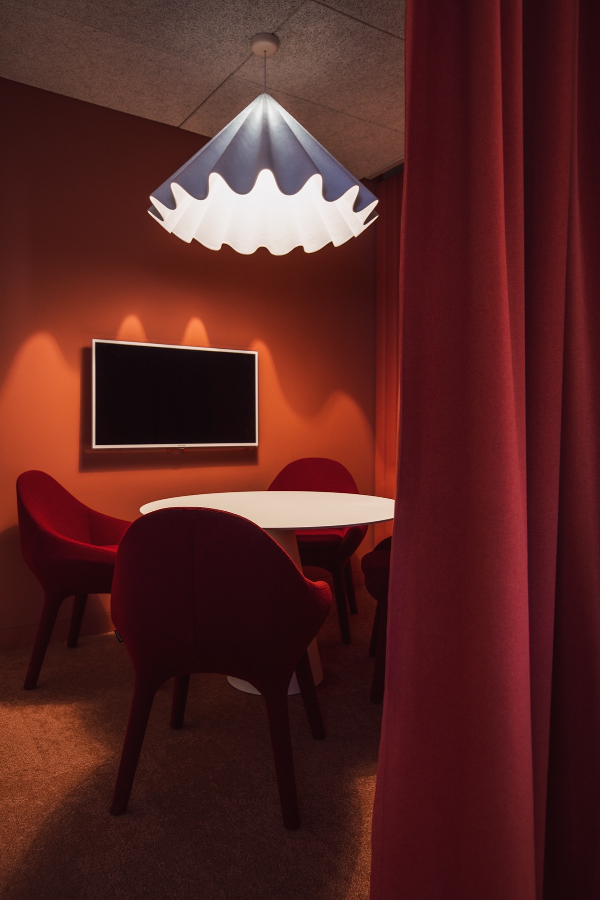 Pomarańczowe biuro z okrągłym białym stołem otoczonym czerwonymi krzesłami, oświetlone designerską lampą i zasłonięte czerwoną zasłoną