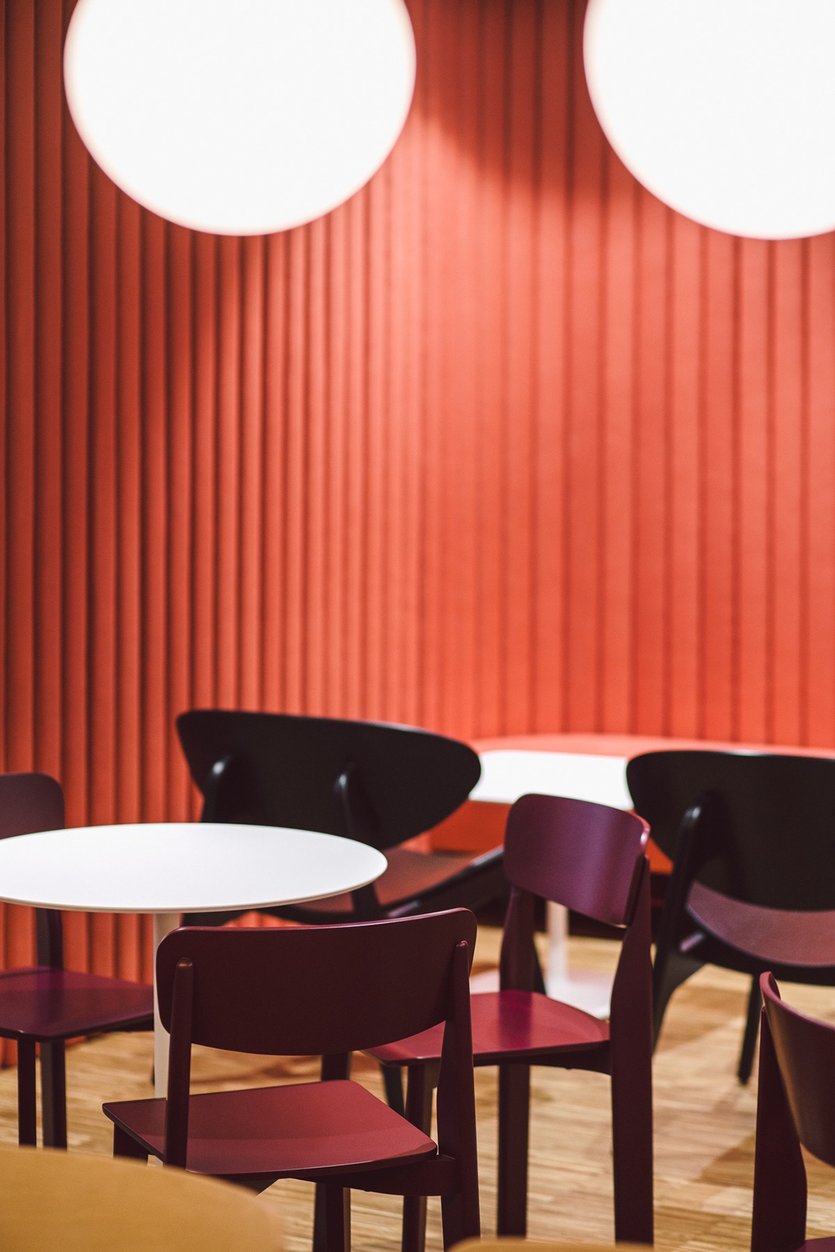 Wnętrze biura z czerwonymi panelami ściennymi, białymi okrągłymi stołami i krzesłami w kolorze burgundu i czerni, oświetlone okrągłymi lampami sufitowymi.