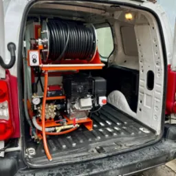 Sprzęt do udrażniania rur zamontowany w przestrzeni ładunkowej białego samochodu dostawczego, widoczny zwój czarnego węża na pomarańczowym stojaku, silnik spalinowy i pompa wysokociśnieniowa.
