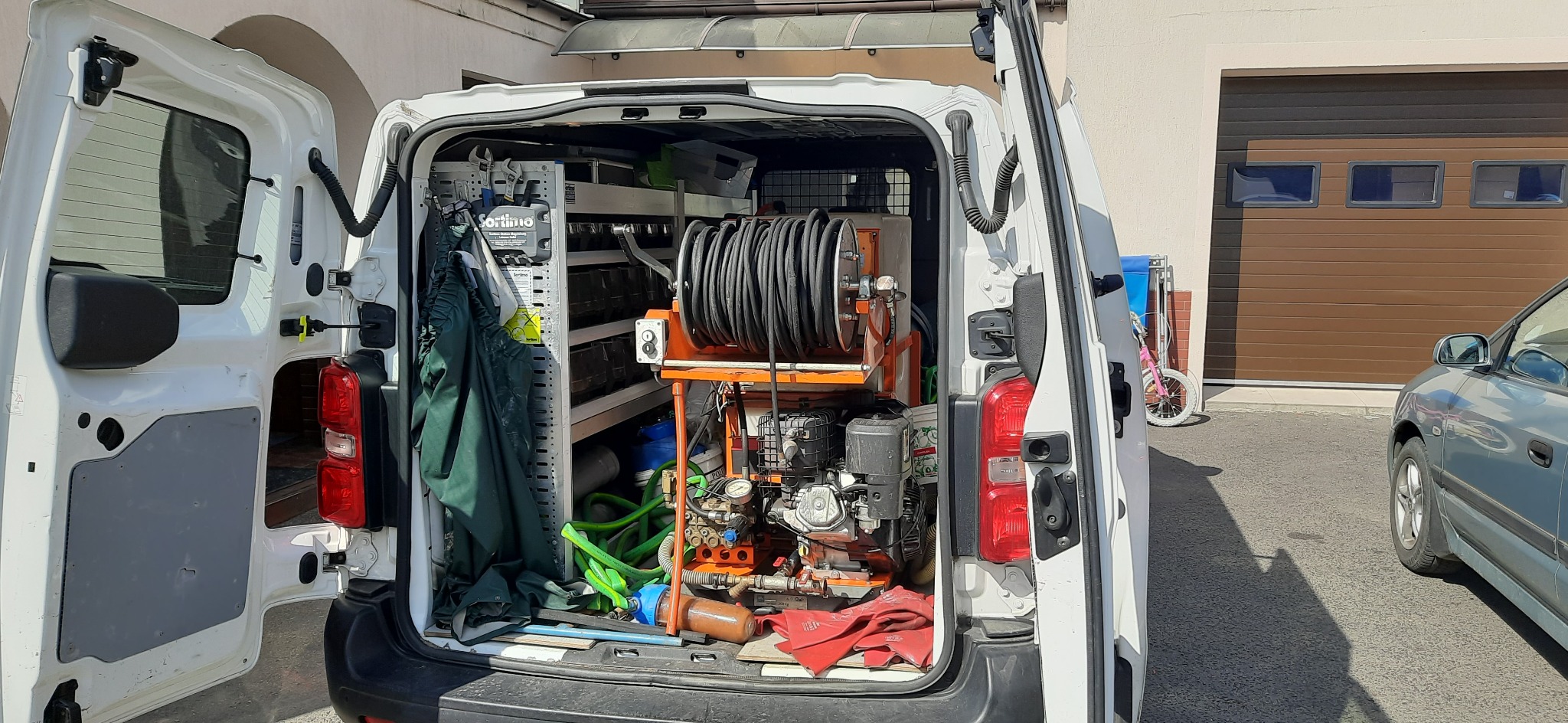 Wnętrze białego samochodu dostawczego z otwartymi drzwiami, widoczny sprzęt do udrażniania rur: zwinięty wąż na pomarańczowym stojaku, agregat ciśnieniowy, narzędzia na półkach; w tle brama...