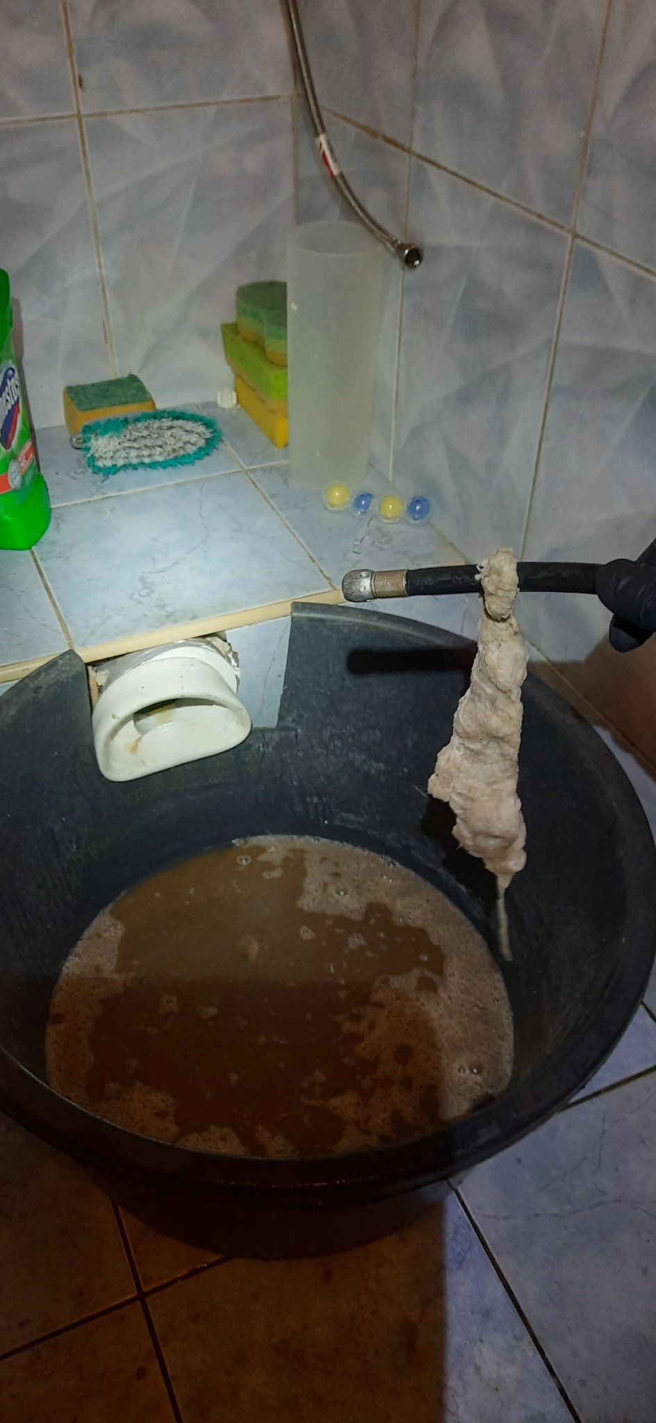 Czarny wąż kanalizacyjny z końcówką zanurzony w wiadrze z brudną wodą, wyciągnięty z rury odpływowej w łazience z niebieskimi płytkami, w tle środki czystości i gąbki.