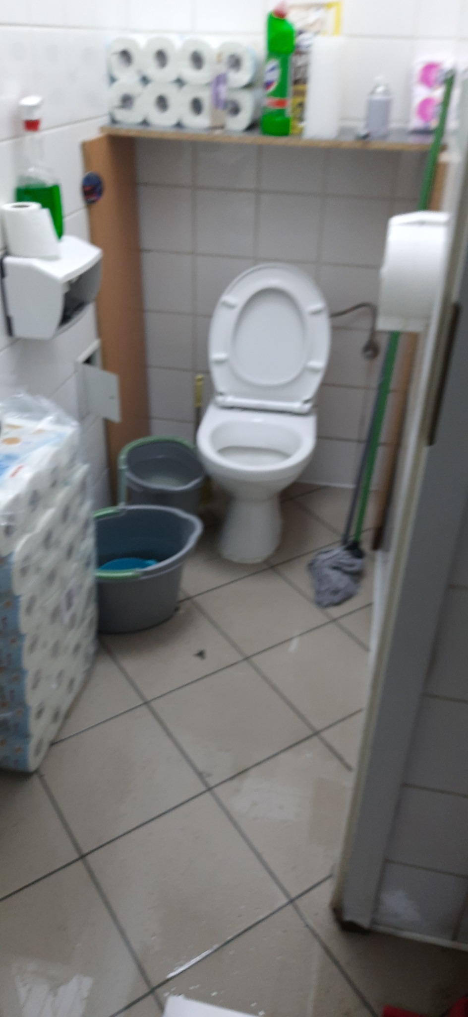 Umywalka z otwartą klapą sedesową, dwa wiadra z wodą, mop i zapas rolek papieru toaletowego w łazience z kwadratowymi płytkami.