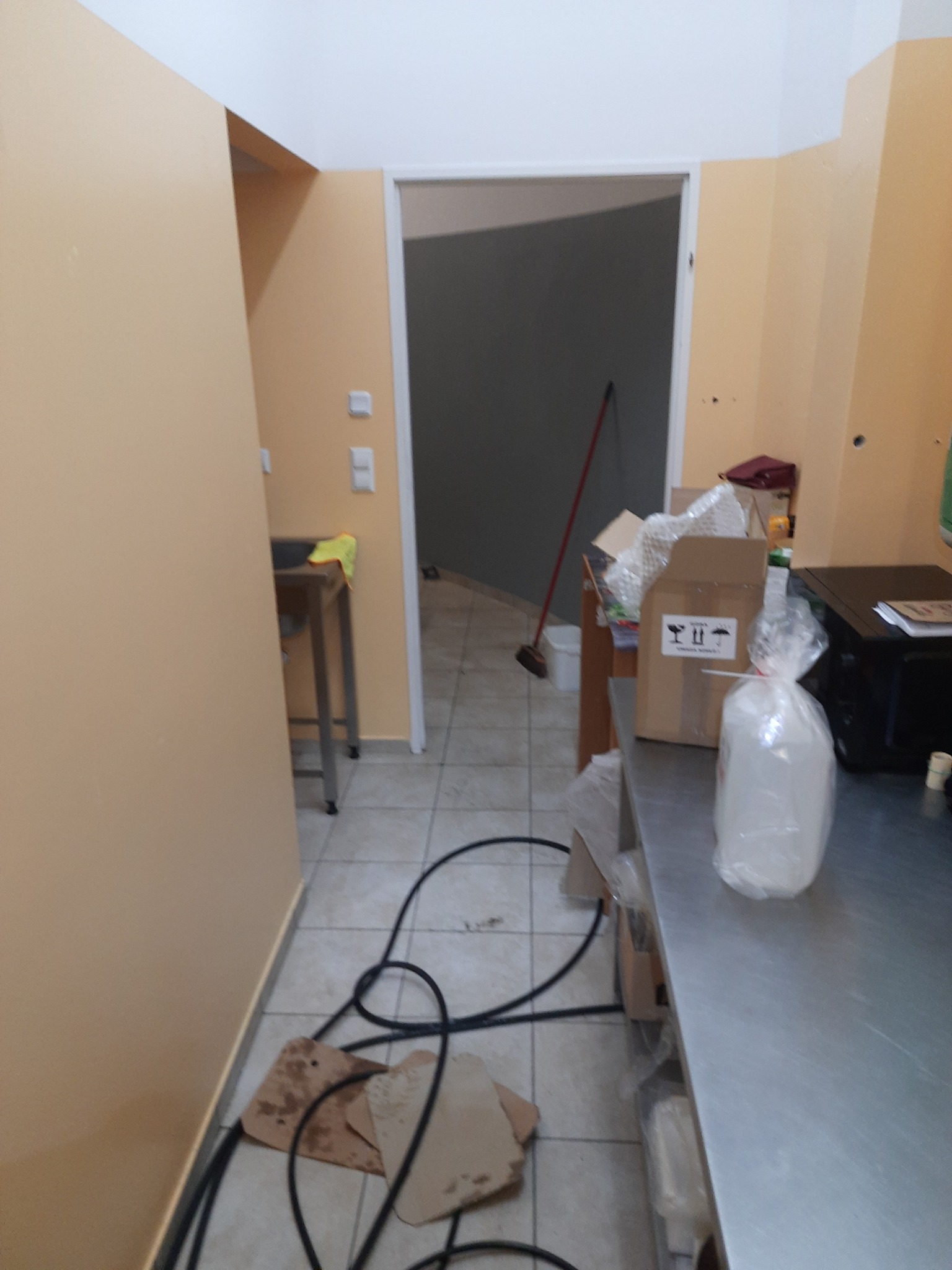 Kable i kartony leżące na podłodze w pomieszczeniu z beżowymi ścianami i kafelkami na podłodze, w tle widoczny otwarty pokój z miotłą.